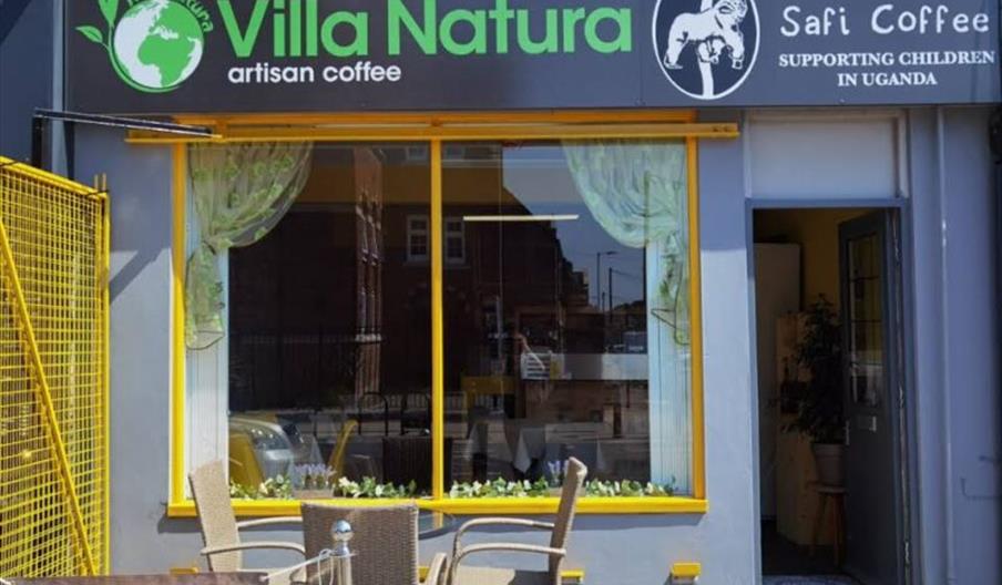 The exterior of Villa Natura, a dark blue coffee shop with yellow edging aroudn the windows. Outside sits a small coffee table with wicker chairs arou