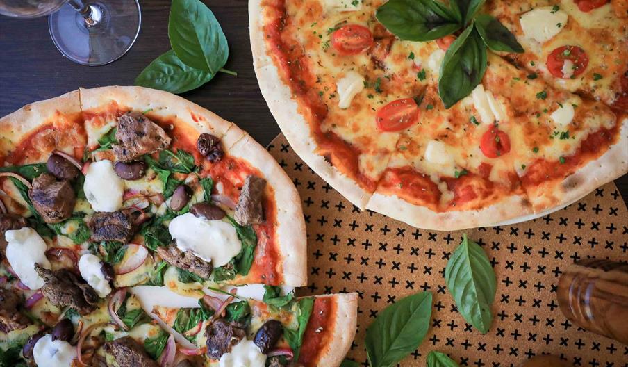 Two pizzas on a table, one topped with cherry tomatoes, cheese, and basil, the other with chunks of meat, olives, spinach, onions, and dollops of chee