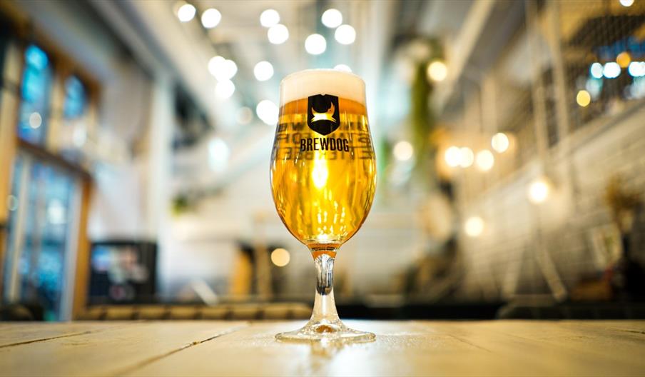 A glass of beer with the BrewDog Logo on, sat on a wooden tabletop with a bar in the background.
