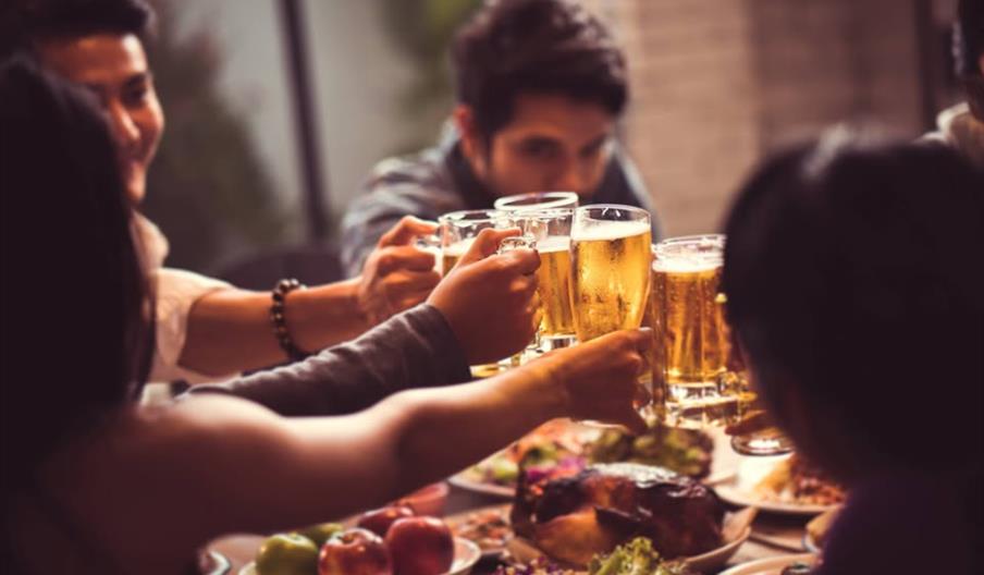 A group of people sitting around a table raise glasses of beer in a toast, with plates of food like fruit and roasted meat in front of them.