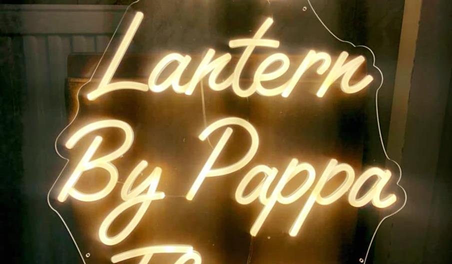 A glowing neon sign in cursive script reads "The Lantern By Pappa Florou" against a dark background.