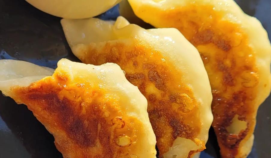 Three golden-brown pan-fried dumplings are arranged on a dark plate next to a small bowl of soy sauce for dipping.