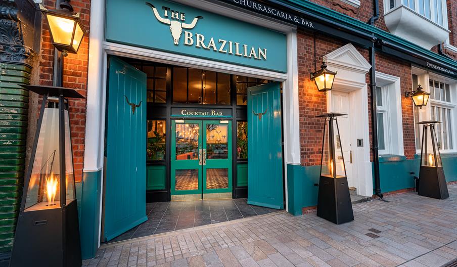 The image shows the entrance to a restaurant called "The Brazilian Churrascaria & Bar," featuring teal double doors, a sign with a bull skull, and out