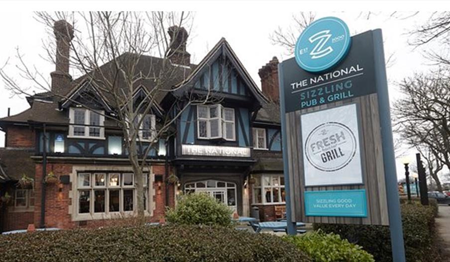 A traditional-style brick and timber pub with large windows and a peaked roof stands behind a prominent sign reading "The National: Sizzling Pub & Gri