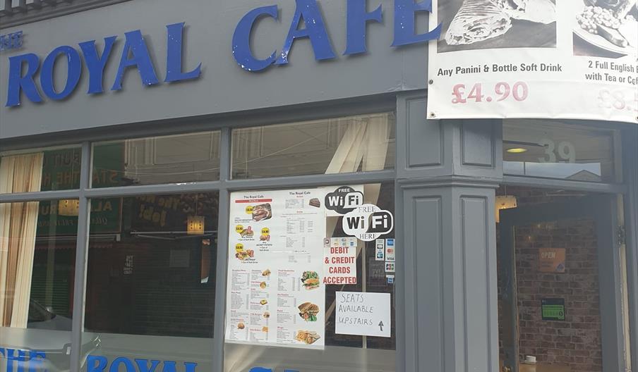 The exterior of The Royal Cafe, with large blue letters on a gray facade. There are multiple signs on the window advertising free WiFi, acceptance of