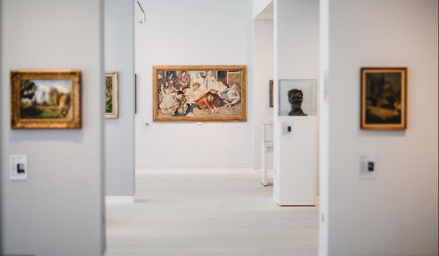 An art gallery with white walls, with assorted paintings hung on them and a sculpture of a head in a glass box off to one side.