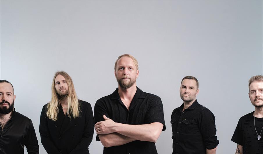 Five men TEMIC wearing black clothing stand in a line against a plain light gray background, with the man in the center crossing his arms and the othe