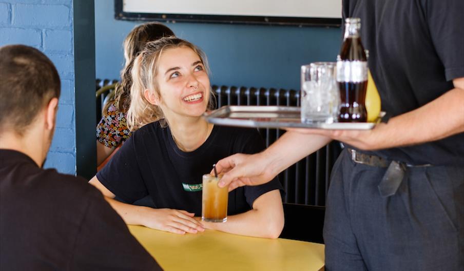 Interior of Thieving Harry's. A server is handing a drink to a smiling woman seated at a table with another person in a café, with a sign reading "m&m