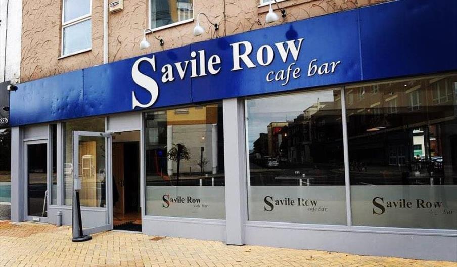 Exterior of Savile Row cafe bar, featuring blue signage and large windows, with a welcoming entrance and tiled pathway.