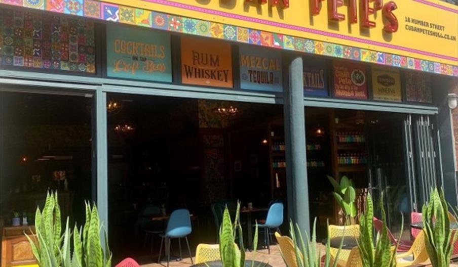 Exterior of red brick building with large open windows , yellow sign with Cuban Petes in red along with colourful tables and chairs on the patio surro