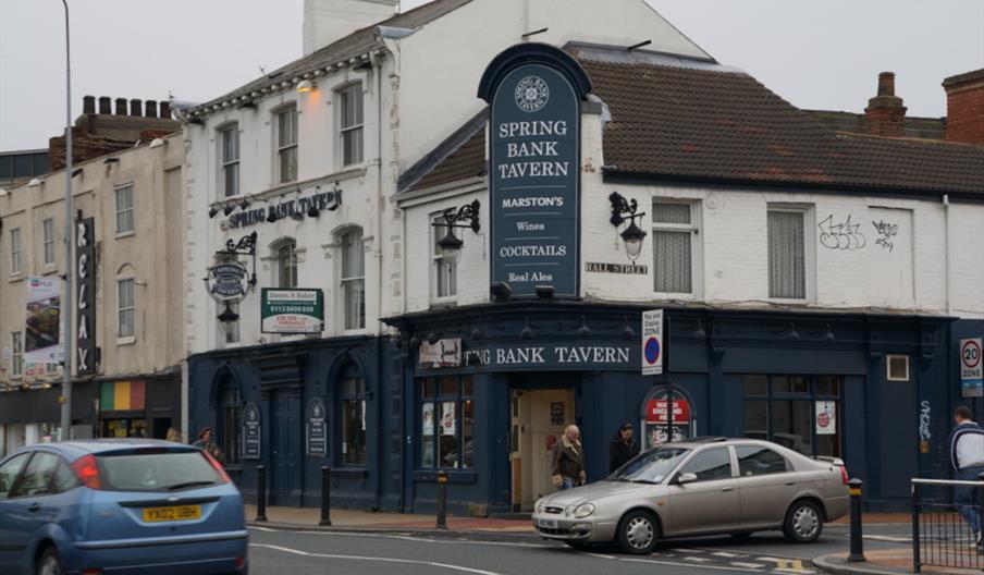 A street view of the Spring Bank Tavern, a traditional pub with a dark blue facade and large sign above the entrance. Several people are walking by an