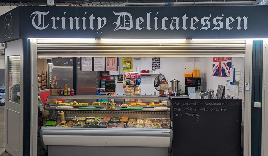 A delicatessen shop named "Trinity Delicatessen" features a refrigerated display case filled with various meats, cheeses, and prepared foods. To the r