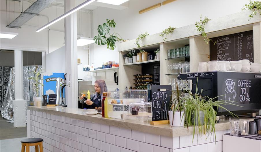 Interior of Humber Street Gallery Cafe.  A modern coffee shop counter with white subway tiles, potted plants, and handwritten chalkboard menus; a bari
