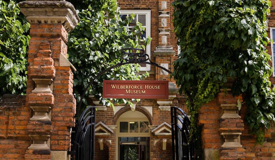 The front gates to the Wilberforce House Museum, with red brick walls either side and foilage spilling over the sides. A hallway leads further into th