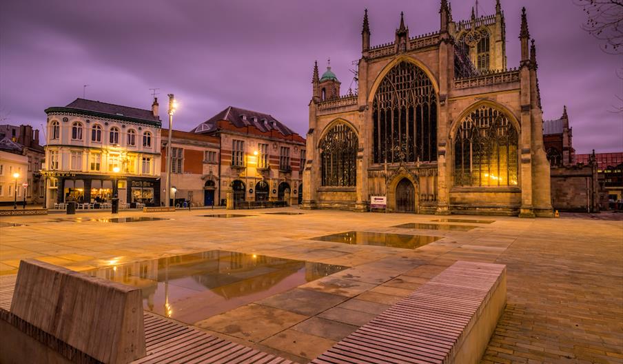 A nightime image of Hull Minster, a large gothic style Minster with stained glass windows and spires. In front there is a wide open paved area sports