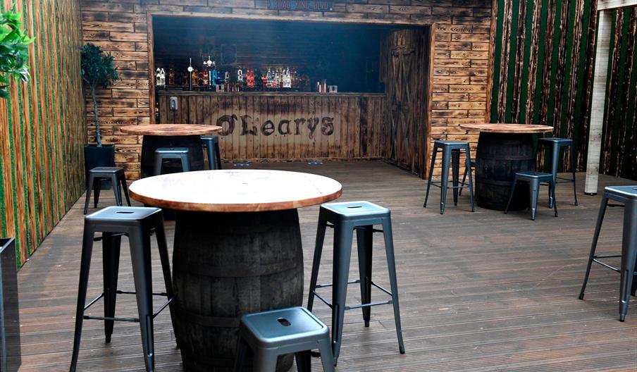A rustic bar area with wooden walls, a wooden countertop labeled "O'Learys," several round tables made from barrels, and black metal stools arranged a