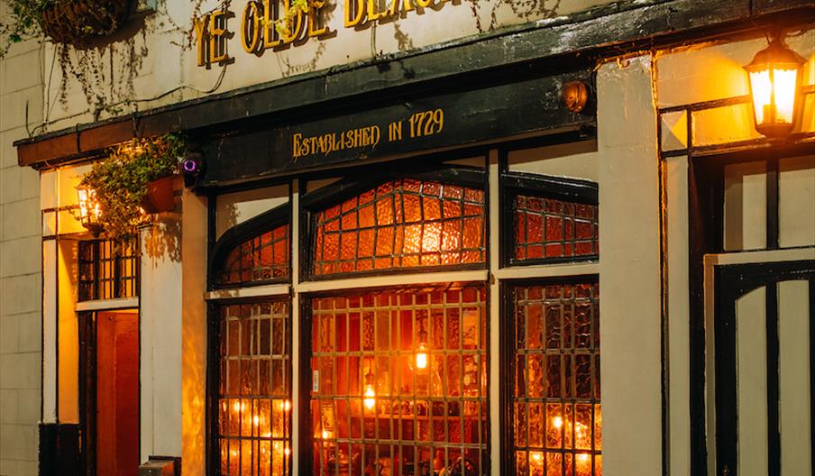 Nighttime street view of "Ye Olde Black Boy" pub with illuminated signage, hanging plants, and vintage-style windows and doors.