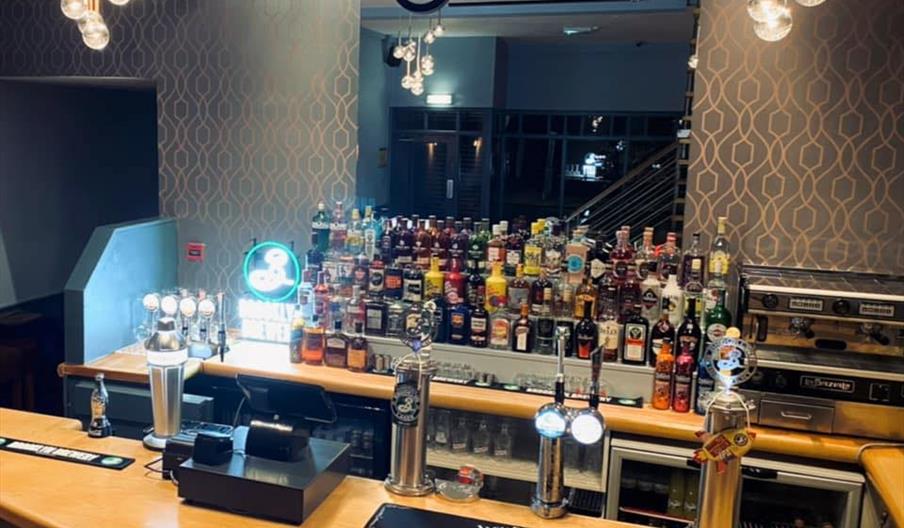 A well-lit bar with a modern design, featuring a curved wooden counter, several beer taps, and a wide selection of liquor bottles displayed on shelves