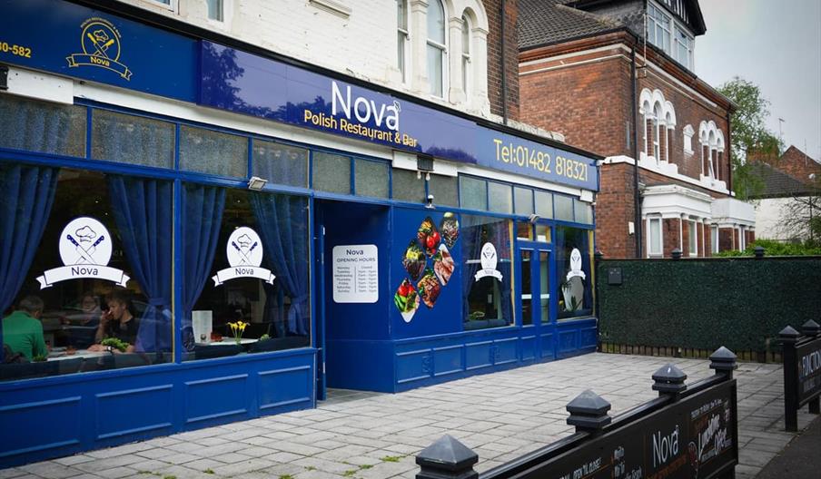 Street view of Nova Polish Restaurant & Bar, a blue-fronted building with large windows, blue curtains, and the restaurant's logo displayed. There are