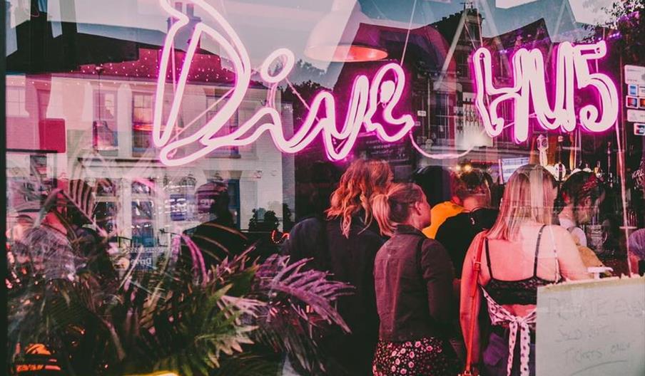 View of bar from outside looking through window with a pink neon sign spelling out Dive HU5 with  a crowd at the bar.