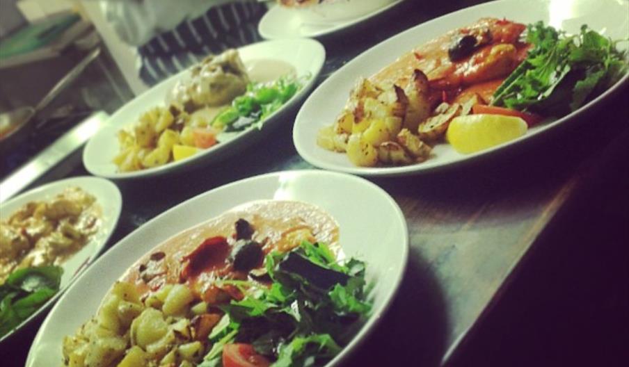 Several plates of food in white bowls on a wooden bench prepared by a chef in a restaurant kitchen.