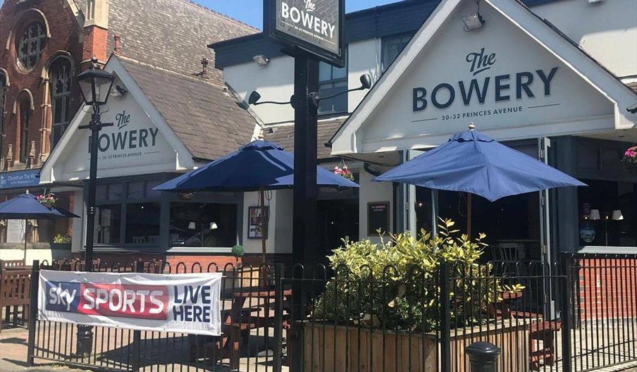 A modern pub called "The Bowery" with large triangular signs, outdoor seating behind a black fence.