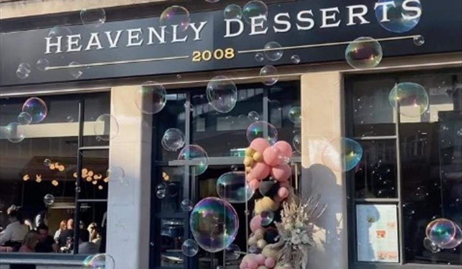 Exterior of "Heavenly Desserts" decorated with a pastel balloon arrangement and a dried floral display. The building features tall windows and columns