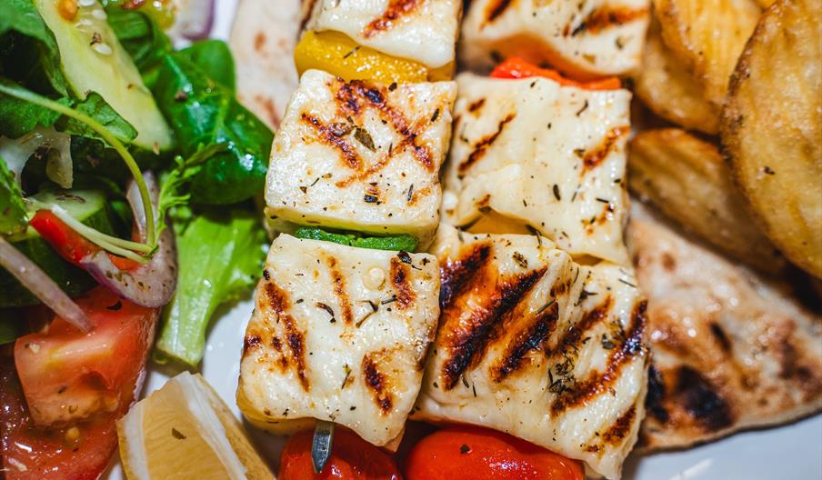 Grilled halloumi cheese skewers with vegetables are served alongside potato wedges, a mixed salad with tomatoes and greens, pita bread, and lemon wedg