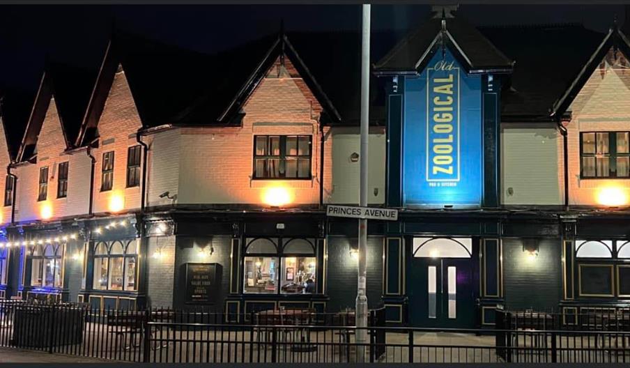 Exterior view of a building at night with a large illuminated sign reading "Old Zoological" above the entrance, and a street sign in front labeled "Pr