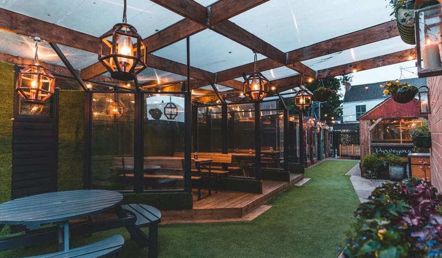 Pubgarden of Larkins Bar. A covered outdoor seating area with wooden beams, large hanging lantern lights, picnic tables, and greenery; some tables are