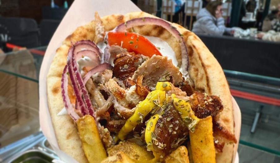 A close-up of a gyro wrapped in pita bread, filled with sliced roasted meat, French fries, red onions, tomato, yellow peppers, and a creamy sauce, hel