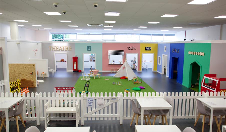 Several playrooms for children around an area with a fake grass in the middle and a small teepee. On the edge of the room is a fence with a seating ar