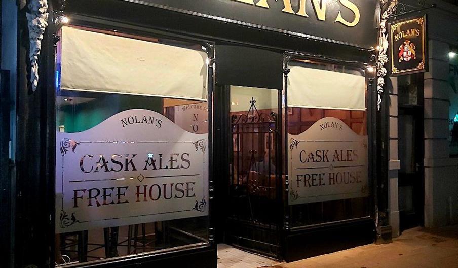 The photo shows the exterior of a pub called "Nolan's" at night, with illuminated gold lettering on a black sign, large front windows displaying etche