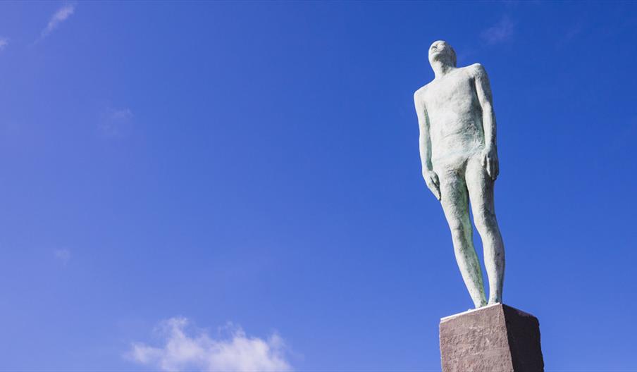 A white statue of a man on a plinth stands against an almost cloudless blue sky.