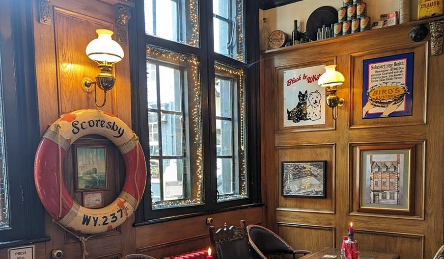 Interior of The Lion and Key. Cozy pub interior with wooden paneled walls, framed artwork, and a large window decorated with string lights; there are
