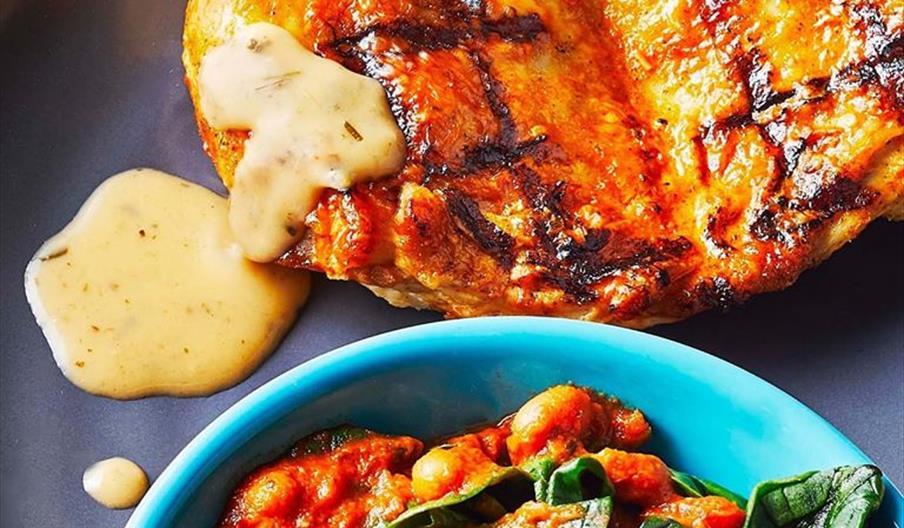 Two grilled chicken breasts with char marks and a creamy sauce are served on a dark plate, alongside a blue bowl filled with stewed tomatoes, white be