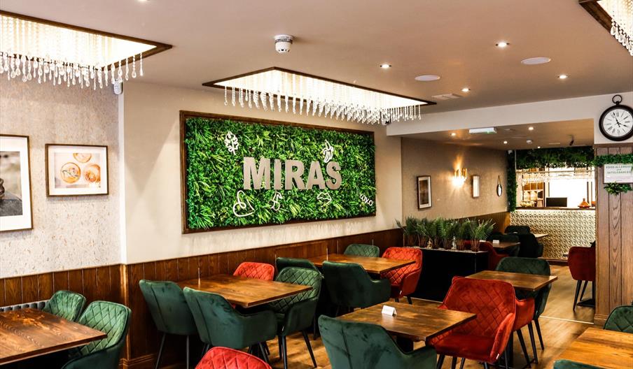 A modern restaurant interior with wooden tables, green and red upholstered chairs, two chandeliers, and a large decorative green wall featuring the wo