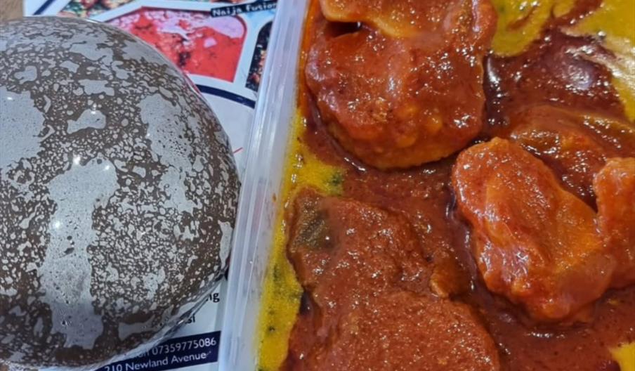 A round serving of dark-colored pounded yam is placed next to a plastic container filled with beef or goat meat covered in red stew, likely a traditio