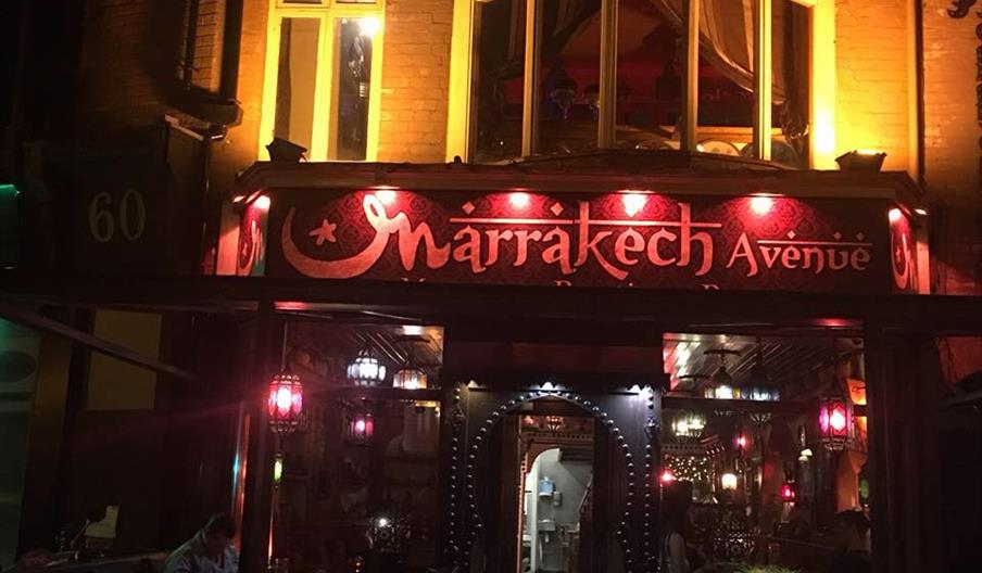 Nighttime view of the entrance to "Marrakech Avenue" restaurant, warmly lit with colorful lanterns, stained glass windows, and decorative signage, cre