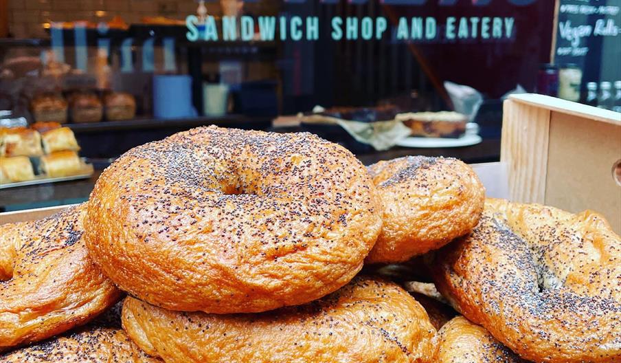 A wooden box filled with poppy seed bagels sits in front of a window displaying the sign "Marla's Sandwich Shop and Eatery."