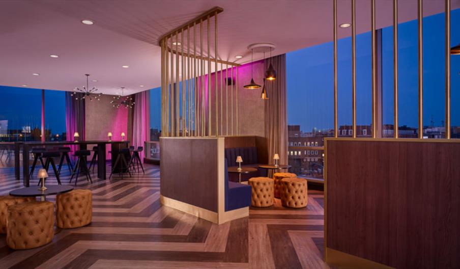 Interior of The Lexington Bar. Modern lounge with large windows showing a cityscape at dusk, featuring patterned wood flooring, blue and gold seating