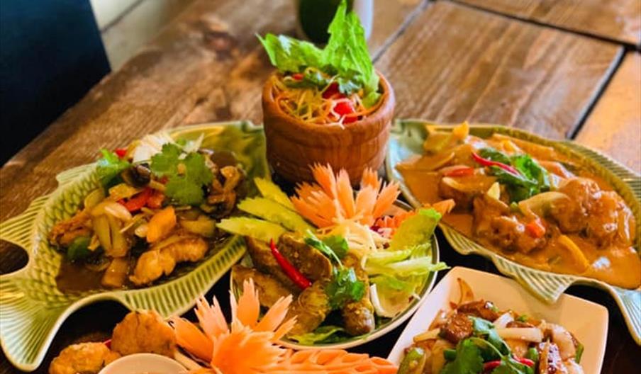 A wooden table set with an assortment of colorful Thai dishes, including stir-fried vegetables with tofu, curry, satay skewers with dipping sauces, sp