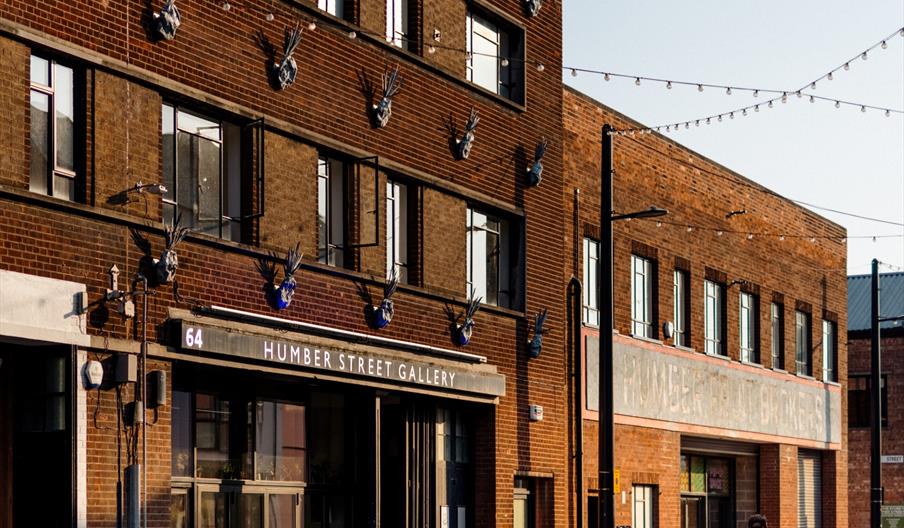 The front of Humber Street Gallery, a three story building witha small seating area out front, several metal pineapples lining the walls and a crowd o