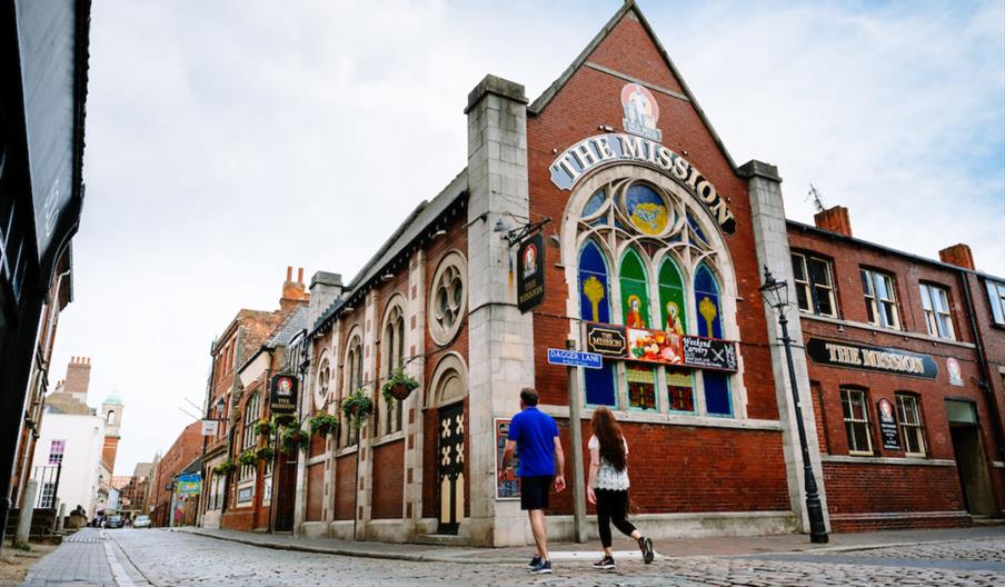 Two people walk down a cobblestone street past a red-brick building with large stained glass windows and a sign reading "The Mission." The building fe