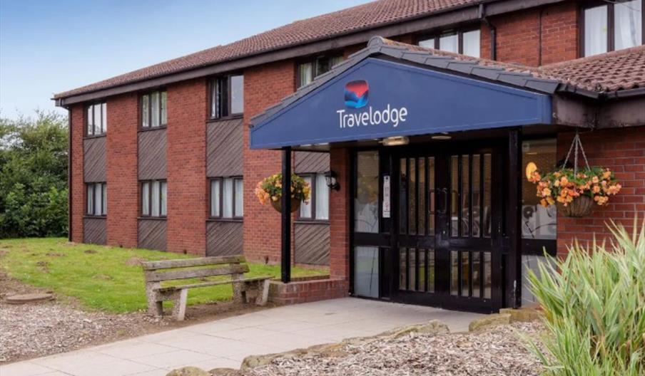 Exterior entrance of a Travelodge hotel, featuring a blue sign with the Travelodge logo above glass double doors, hanging flower baskets on either sid