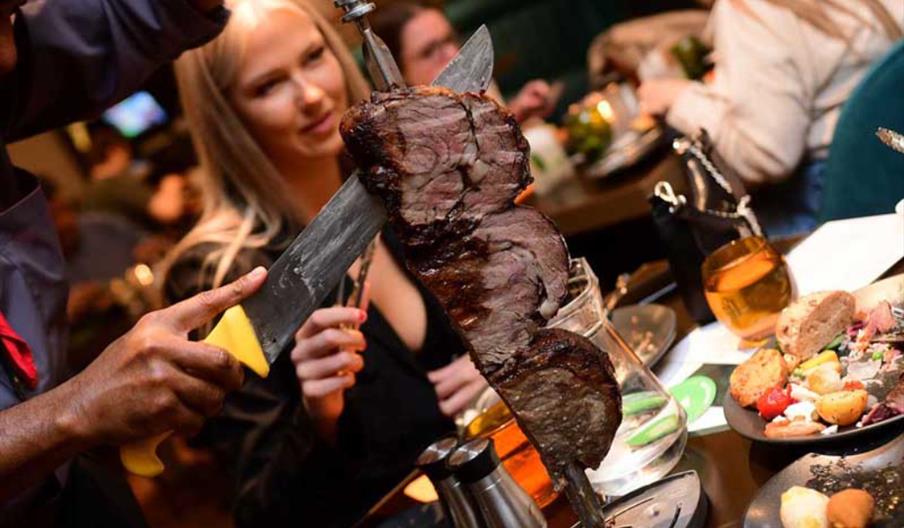 Meat on a large skewer sliced infront of people in a busy restaurant