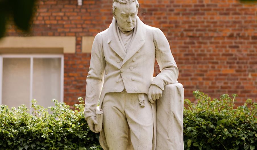 A white statue of William Wilberforce, wearing Edwardian clothing and leaning on a post, in front of a red bricked building with large bay windows.