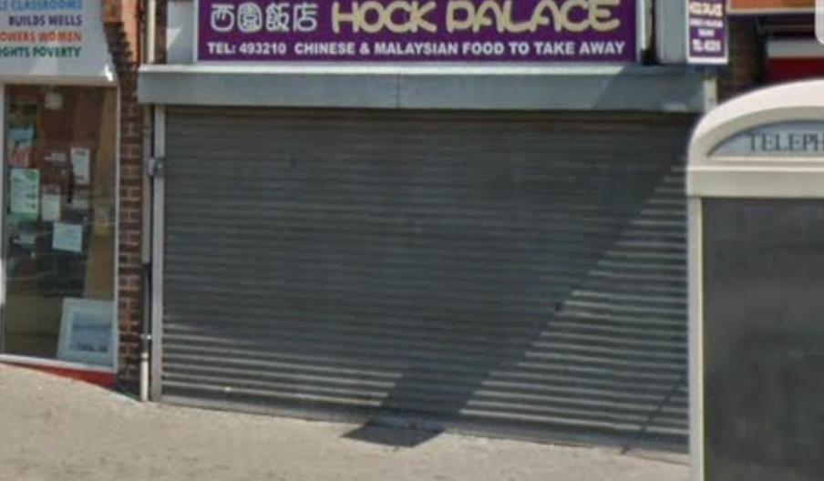 A closed takeaway restaurant with a metal shutter pulled down, featuring a purple sign above that reads "Hock Palace Chinese & Malaysian Food to Take
