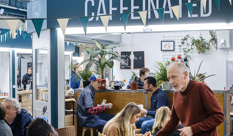A group of people around various coffee tables with a coffee shop operating out of a market unit in the background with the word 'Caffeinated'.
