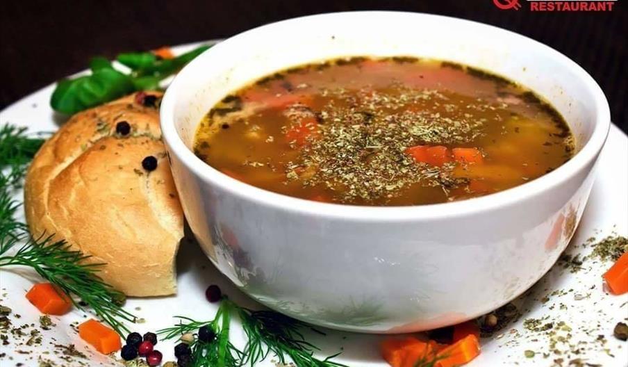 A white bowl filled with vegetable soup, garnished with herbs, sits on a plate next to a round bread roll. Fresh dill, diced carrots, and peppercorns